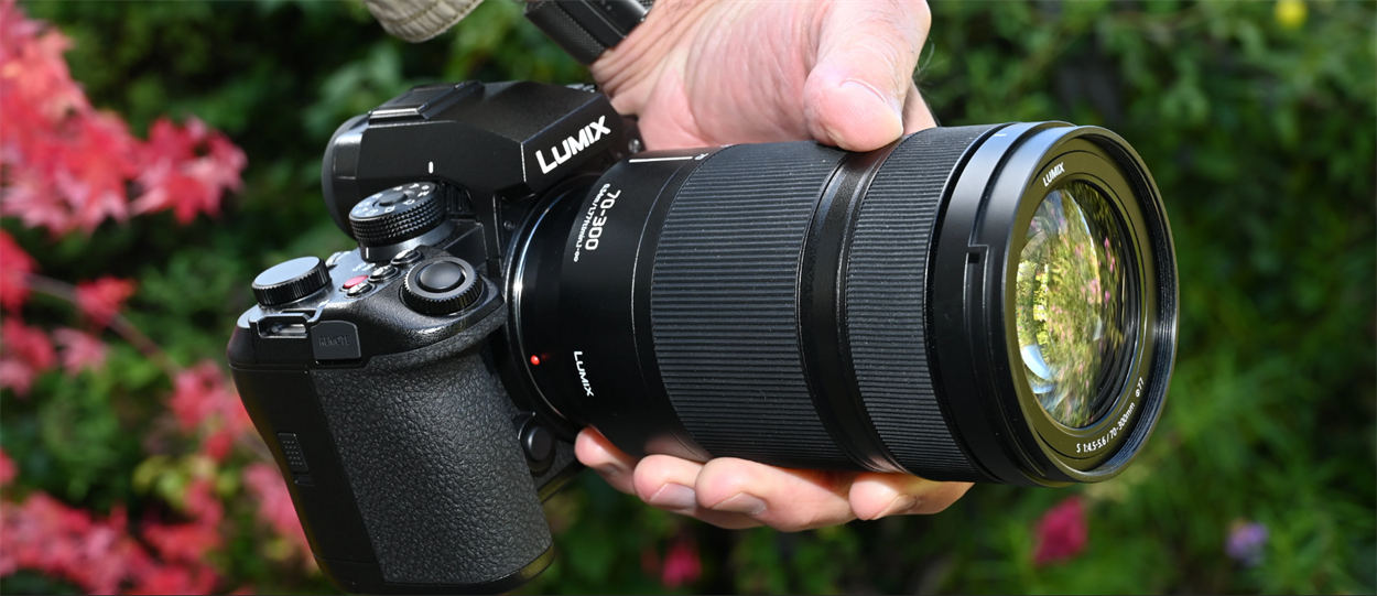 Руки фотографа тримають бездзеркальну камеру Lumix з встановленим телеоб'єктивом Lumix S 70-300mm на тлі зеленого листя та червоних квітів.