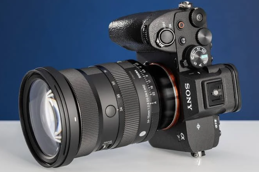Камера Sony серії Alpha з підключеним зум-об'єктивом Sigma 24-70mm f/2.8 DG DN II стоїть на білій поверхні перед синім фоном.